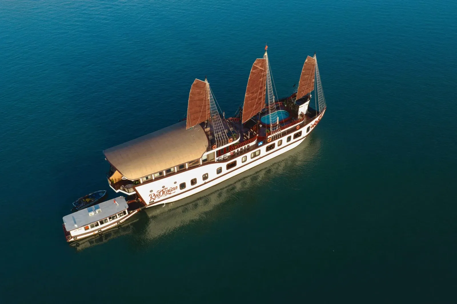 An aerial drone view of a white three-masted junk-style cruise boat with brown sails and the name 'Red Dragon' painted on its hull, with a smaller tender boat and a speedboat alongside on calm dark teal water.