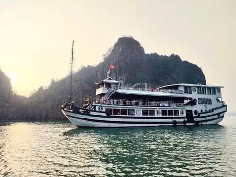 A white and brown cruise boat with multiple decks and a red flag floats on calm green water with limestone karsts in the background.