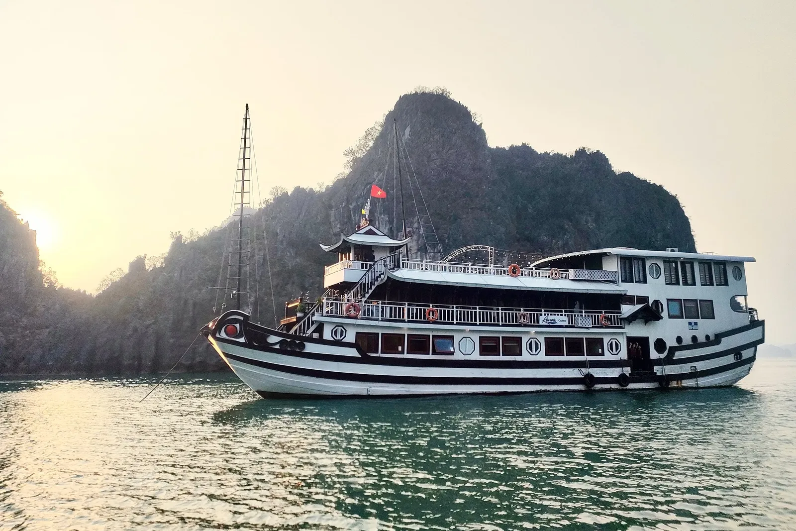A white and brown cruise boat with multiple decks and a red flag floats on calm green water with limestone karsts in the background.