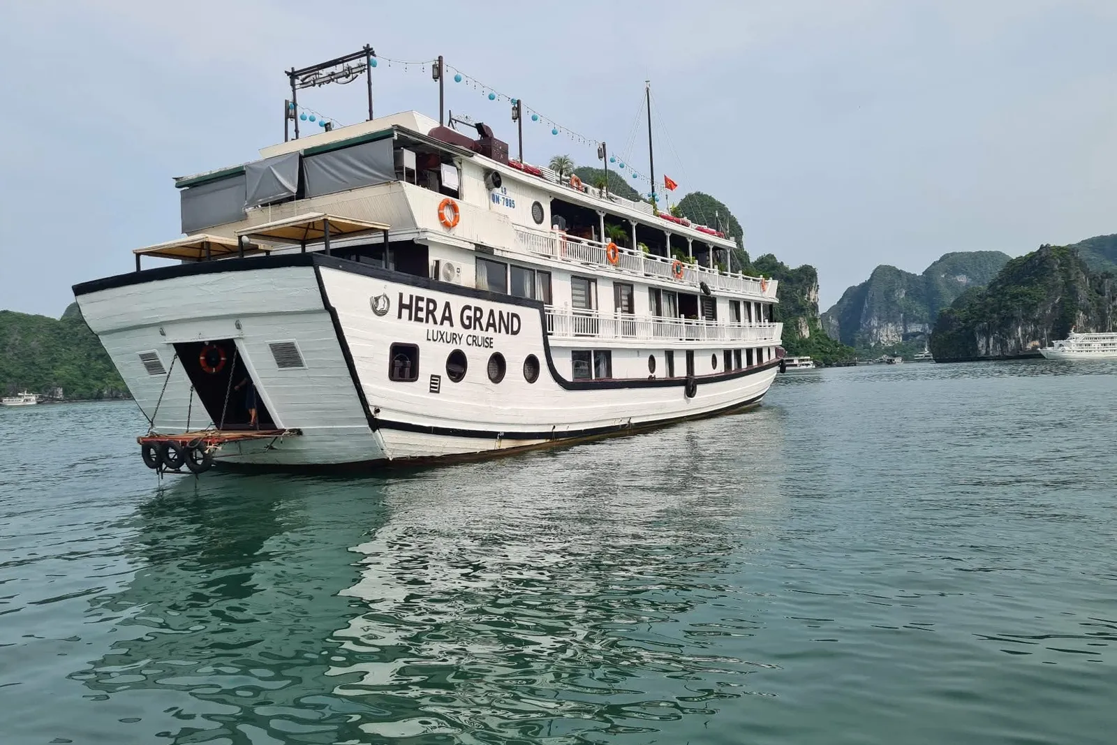 A white multi-deck cruise boat with 'HERA GRAND LUXURY CRUISE' painted on its stern sits on calm green water in front of forested limestone karst islands, with another vessel visible in the distance.