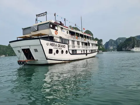 A white multi-deck cruise boat with 'HERA GRAND LUXURY CRUISE' painted on its stern sits on calm green water in front of forested limestone karst islands, with another vessel visible in the distance.