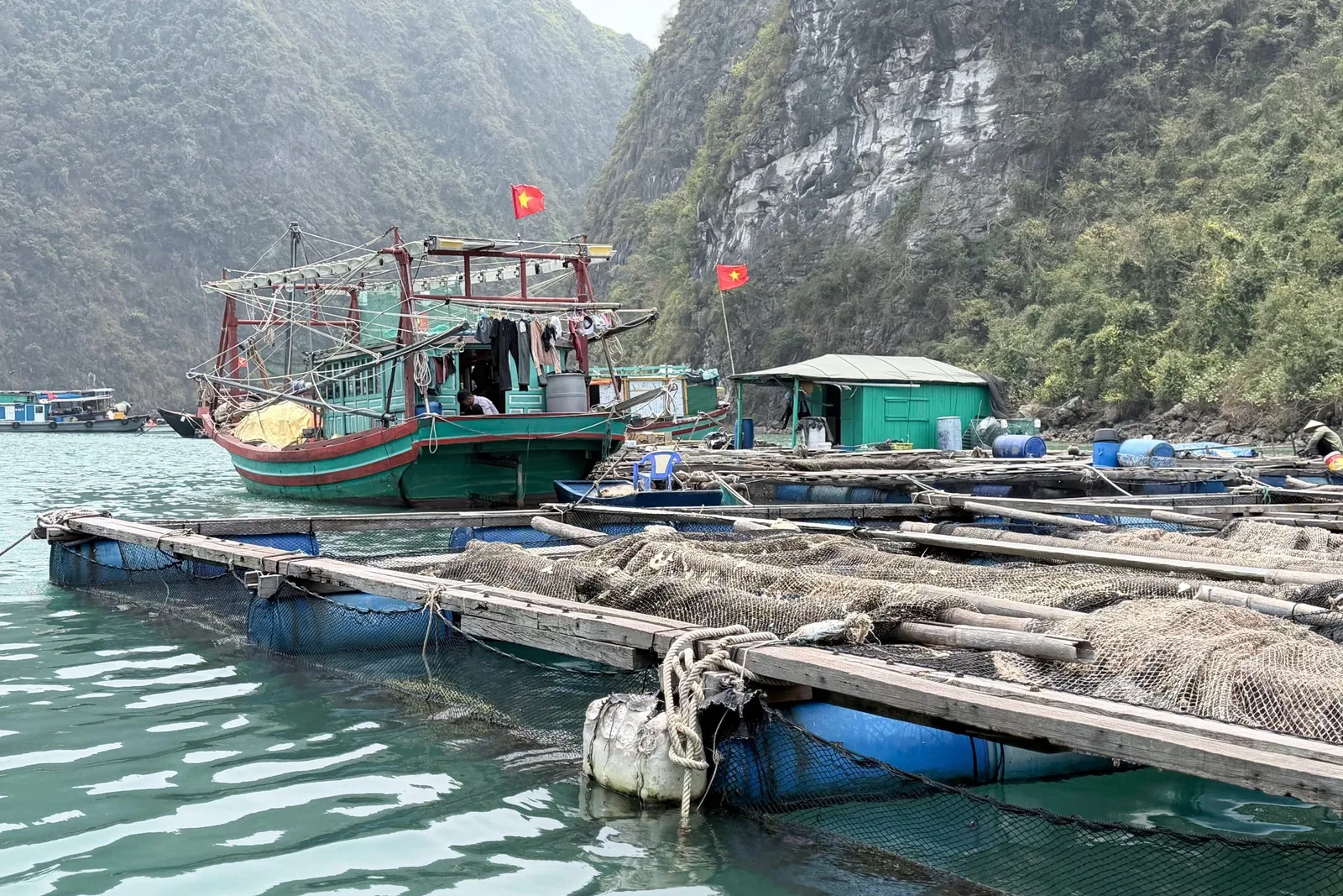 A green and red fishing boat with Vietnamese flags is moored next to floating fish farm platforms surrounded by limestone karst mountains.