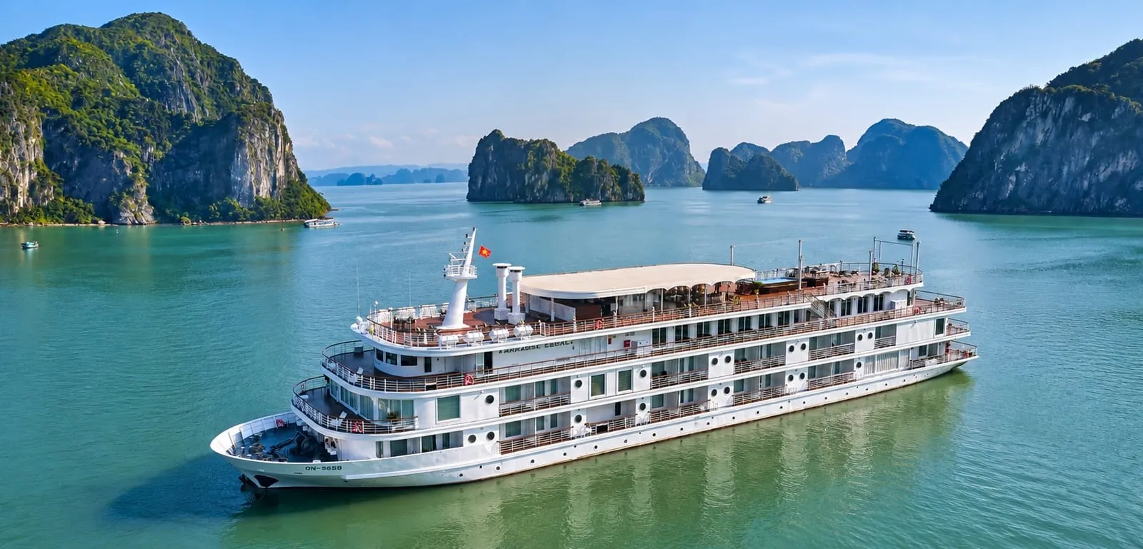 An aerial view of a large white multi-deck cruise ship with a small red flag on green-turquoise water, surrounded by limestone karst islands under a blue sky.