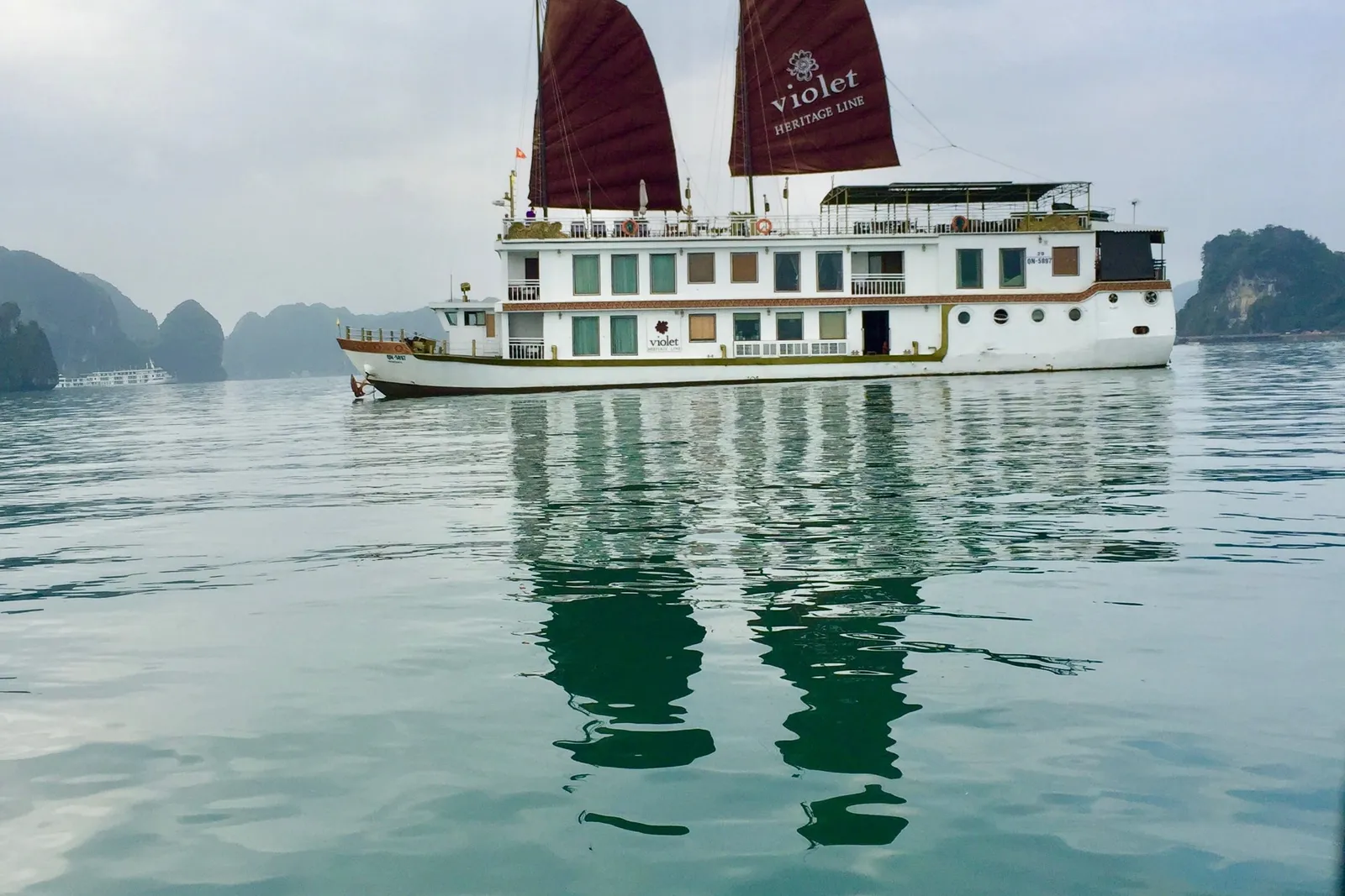 A white two-deck vessel with two dark red sails printed 'violet Heritage Line' sits on calm green water with limestone karsts and a distant boat behind it, seen from another boat.