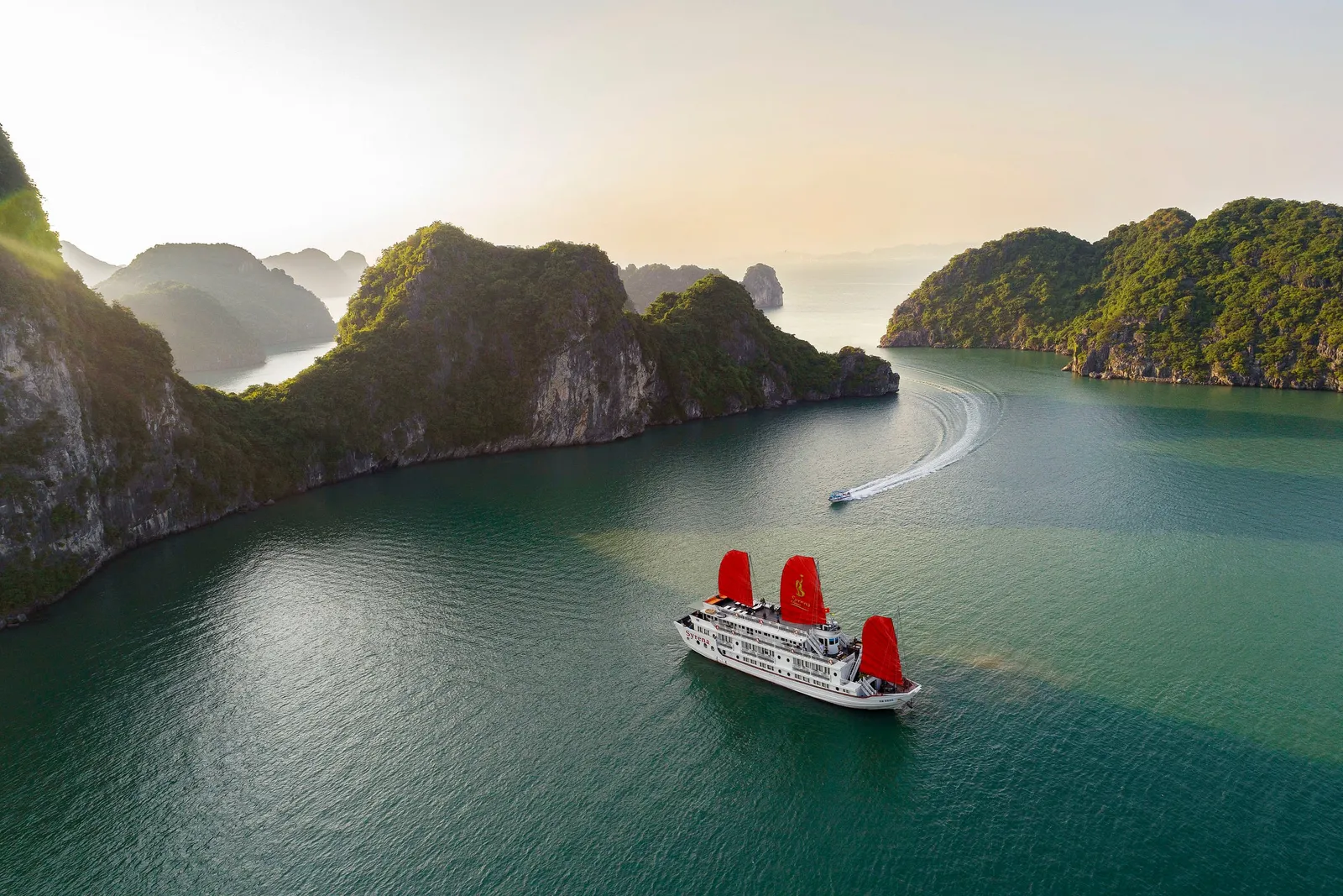 Aerial view of a white cruise ship with three red sails, one carrying a small gold emblem, anchored in calm green water among forested limestone karst islands, with a small speedboat leaving a wake in the background.