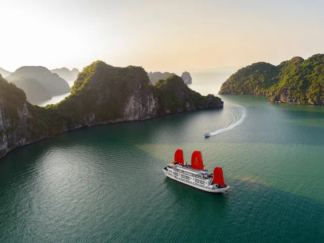 Aerial view of a white cruise ship with three red sails, one carrying a small gold emblem, anchored in calm green water among forested limestone karst islands, with a small speedboat leaving a wake in the background.