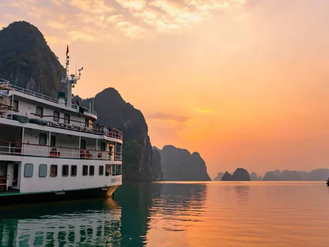 A white multi-deck cruise boat with the name 'Emeraude' on its hull sits on calm water beside limestone karst islands under an orange sunset sky, with a small boat in the distance.