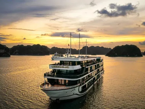 A white multi-deck cruise ship with 'LA REGINA GRAND' lettered on its upper deck, seen from an elevated aerial angle on calm water at sunset, with limestone karst islands and a golden sky behind it.