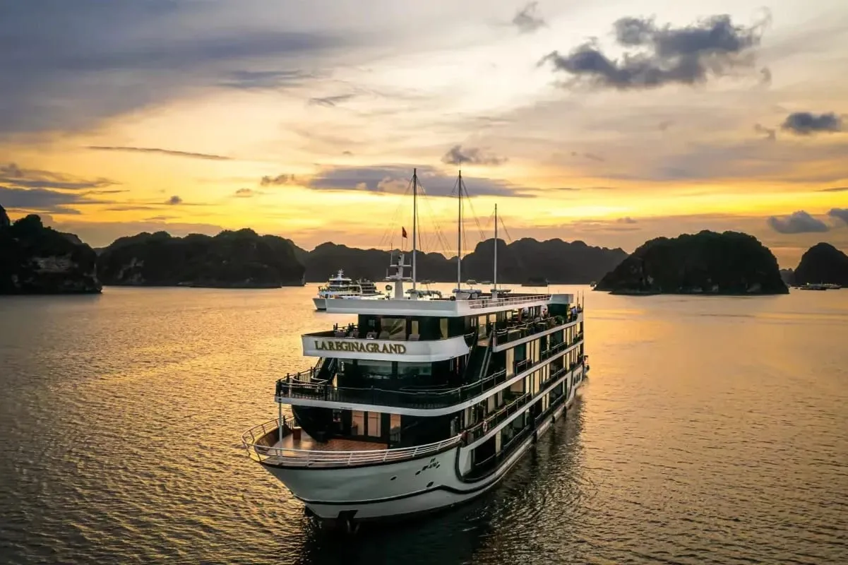 A white multi-deck cruise ship with 'LA REGINA GRAND' lettered on its upper deck, seen from an elevated aerial angle on calm water at sunset, with limestone karst islands and a golden sky behind it.