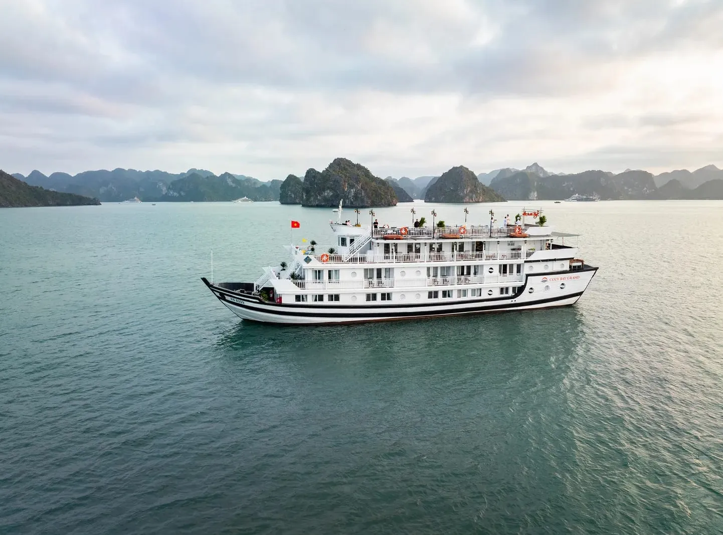 Aerial view of a white multi-deck cruise ship with a black stripe and a Vietnamese flag on calm green water, with limestone karst islands in the background and small lettering on the hull.