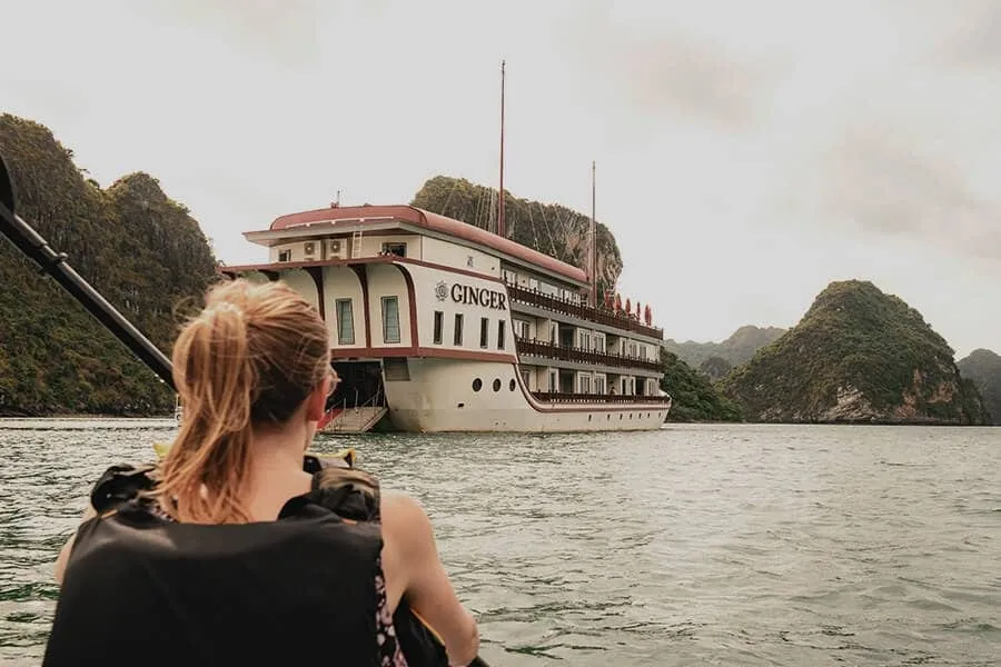A woman in a black life vest with a ponytail sits in a kayak facing a multi-deck white and maroon cruise ship with 'GINGER' on its hull, surrounded by green limestone karst islands on calm water.