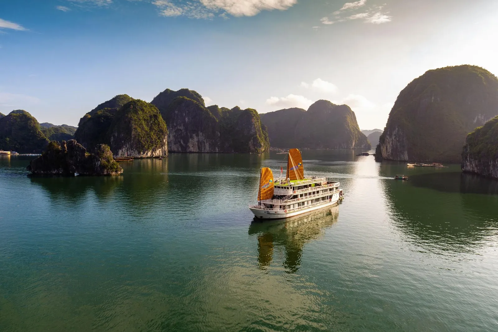An aerial view of a multi-deck white cruise boat with orange sails on calm green water, surrounded by forested limestone karst islands under a blue sky with clouds.
