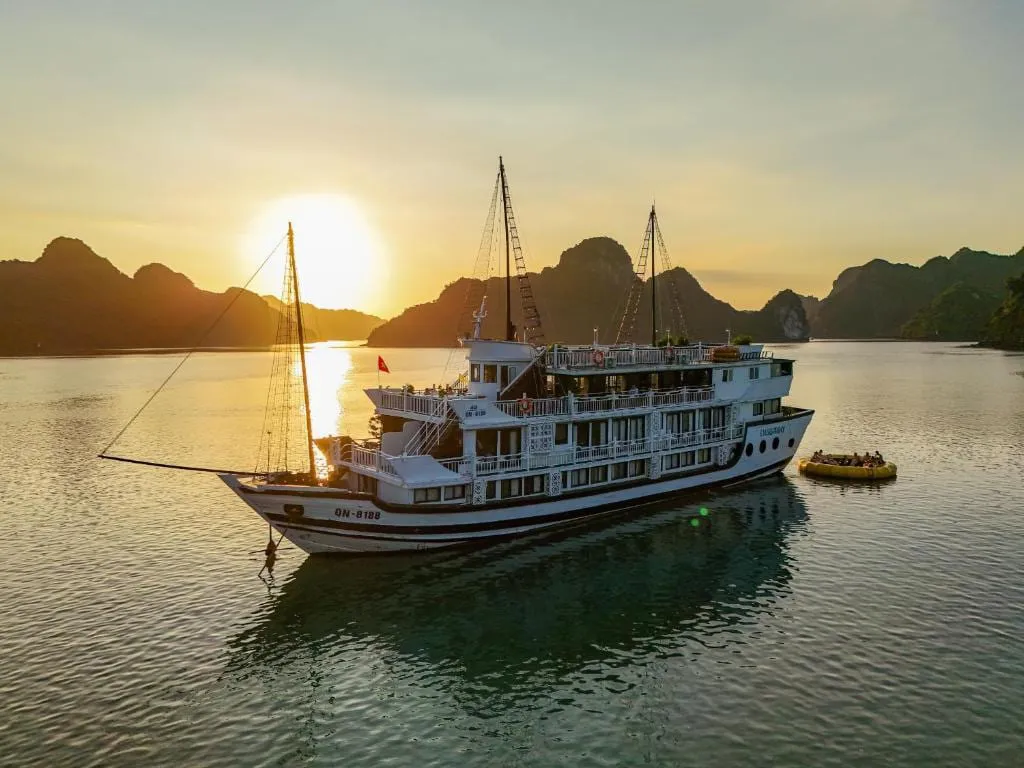 An aerial view of a white multi-deck wooden cruise boat with masts, registration number QN-8188 on the bow and a small name on the hull, anchored in calm water at sunset with limestone karst islands behind and a yellow inflatable raft with people nearby.