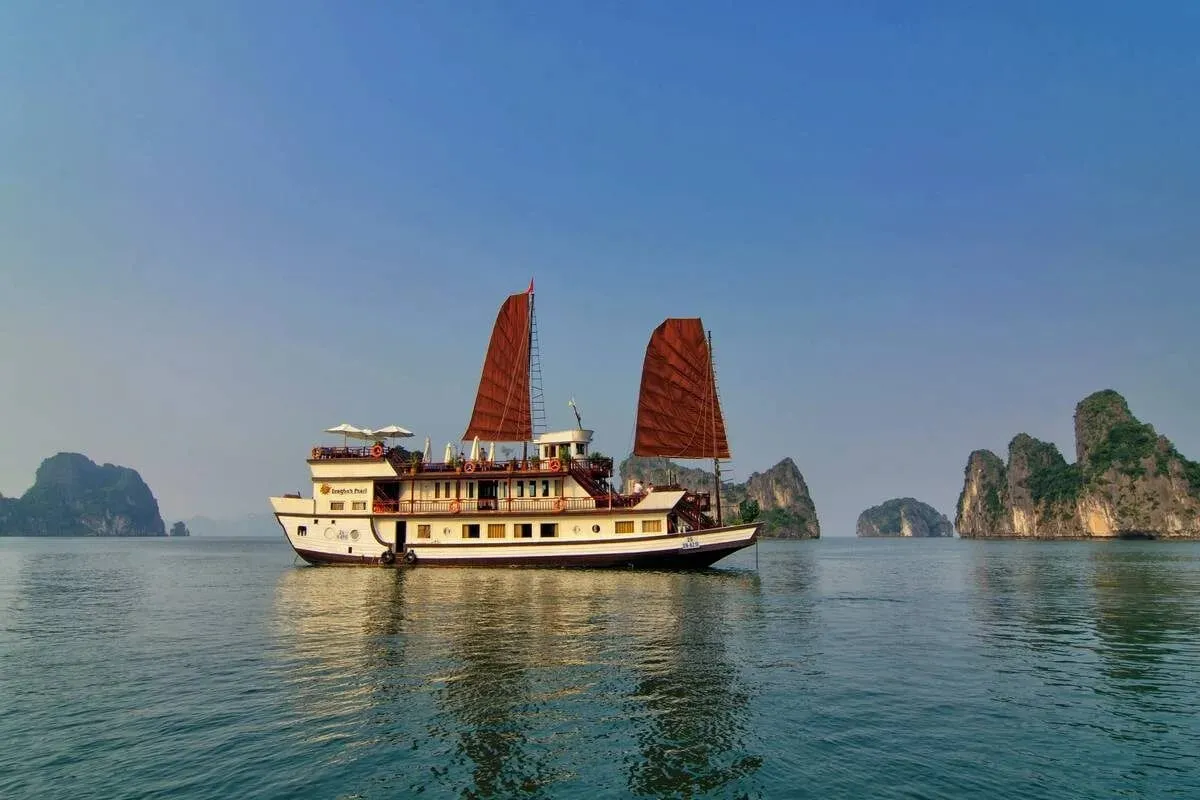 A cream wooden junk with two red sails in a three-quarter view with limestone islands behind it.