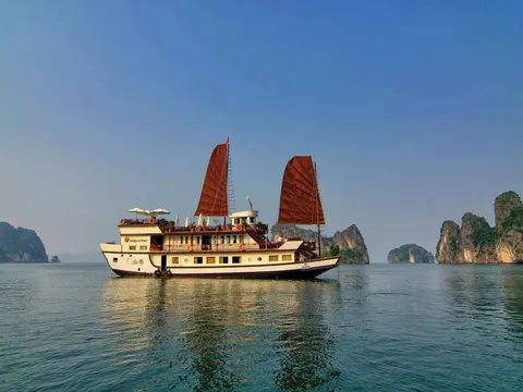 A cream wooden junk with two red sails in a three-quarter view with limestone islands behind it.