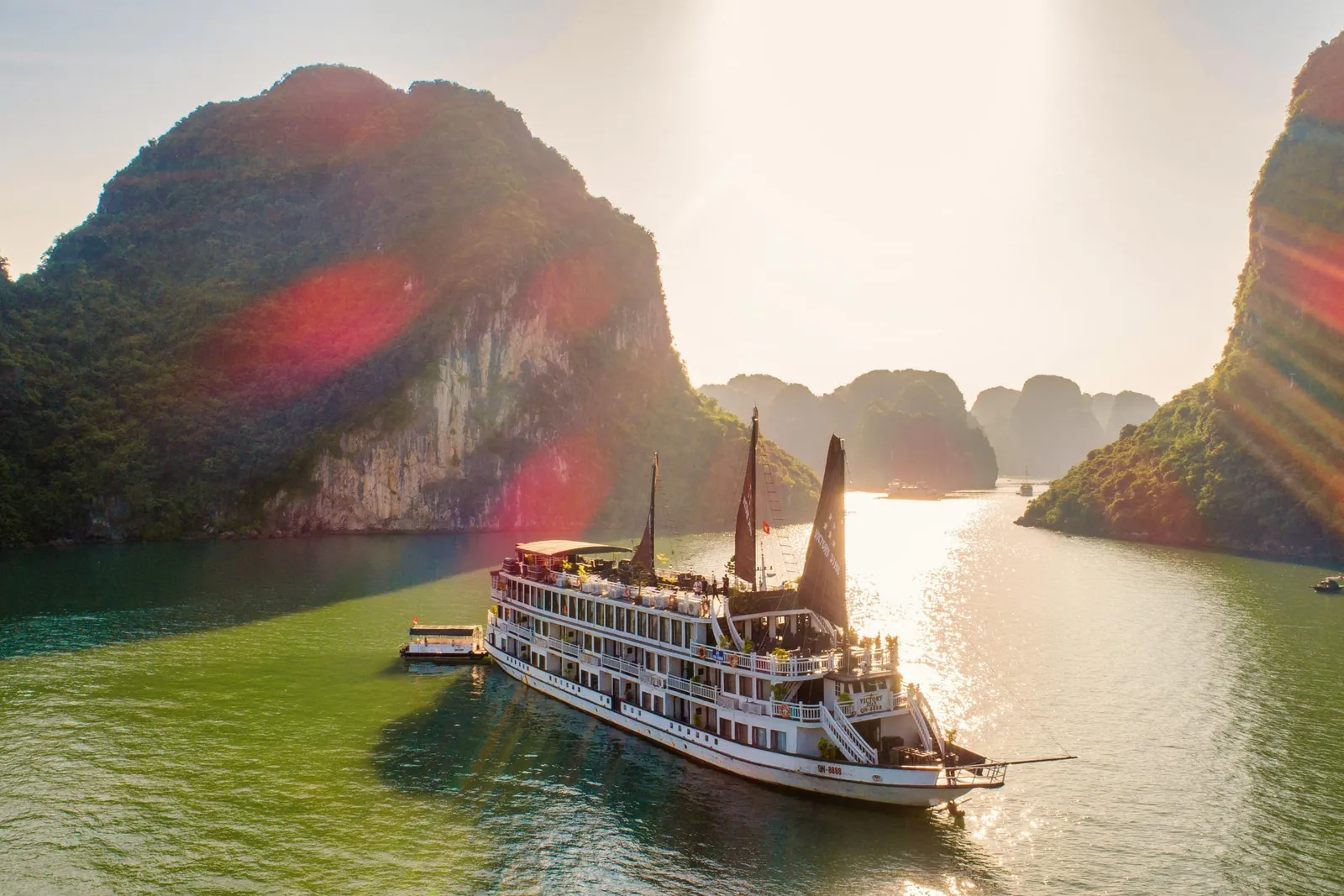 A large white cruise ship with traditional sails floats in calm green water surrounded by tall limestone karst islands at sunset.