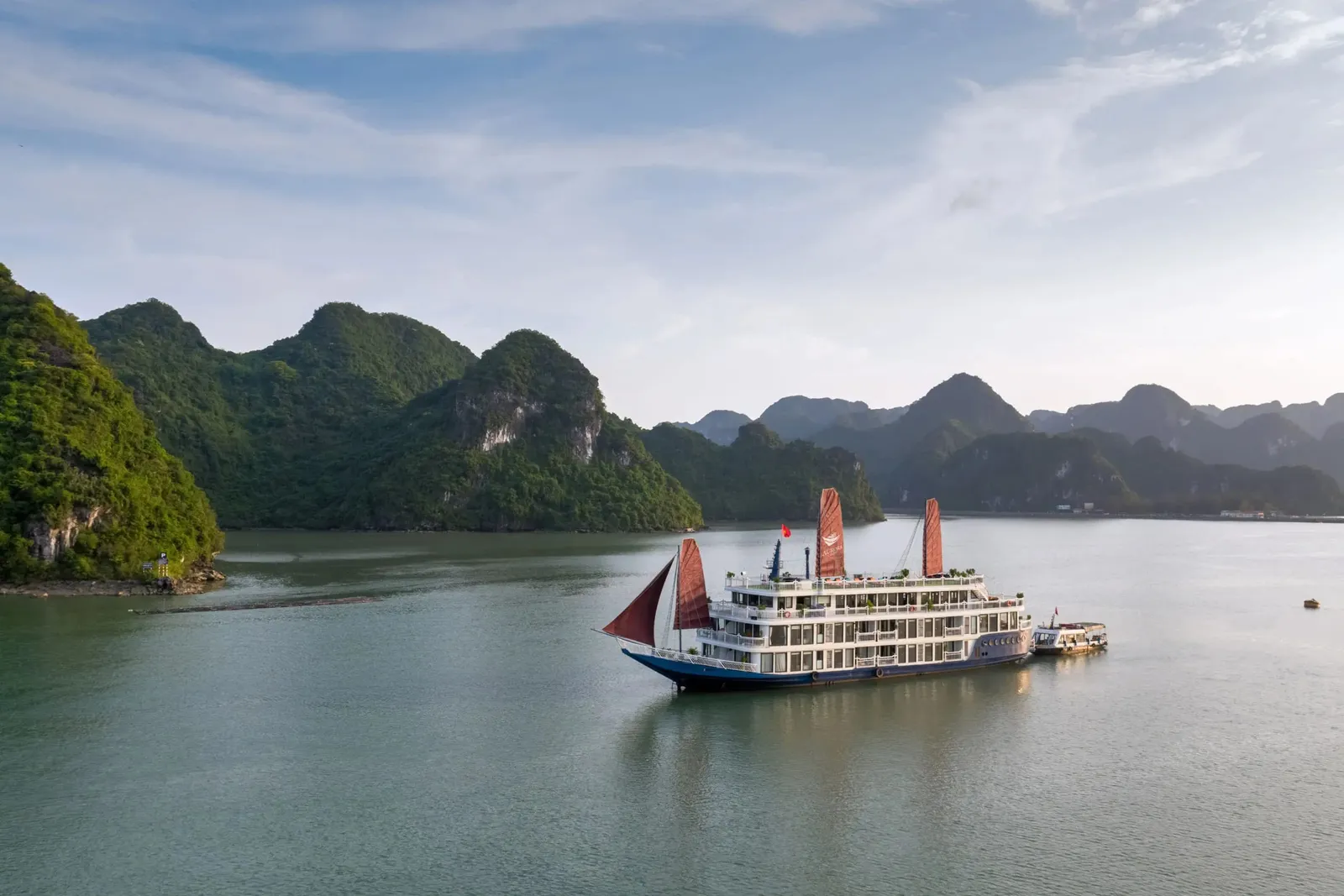 A multi-deck white cruise boat with a blue hull and red-orange sails sits on calm green water, with a small tender boat beside it and forested limestone karst islands in the background under a soft blue sky.