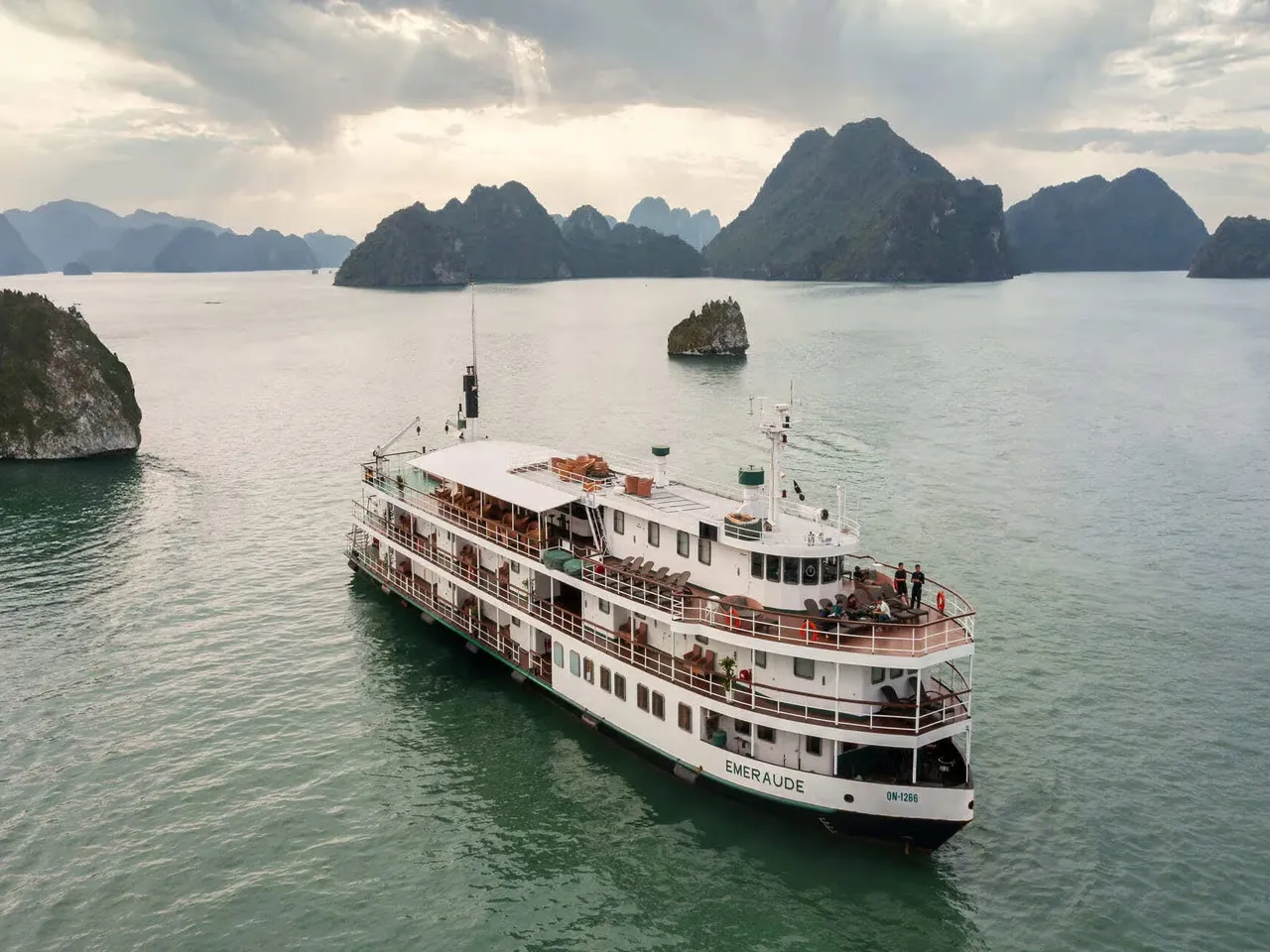 Aerial view of a white multi-deck cruise ship with 'EMERAUDE' on its stern, anchored in green water among limestone karst islands under a cloudy sky.