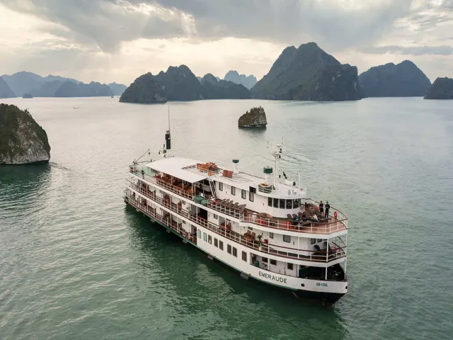 Aerial view of a white multi-deck cruise ship with 'EMERAUDE' on its stern, anchored in green water among limestone karst islands under a cloudy sky.