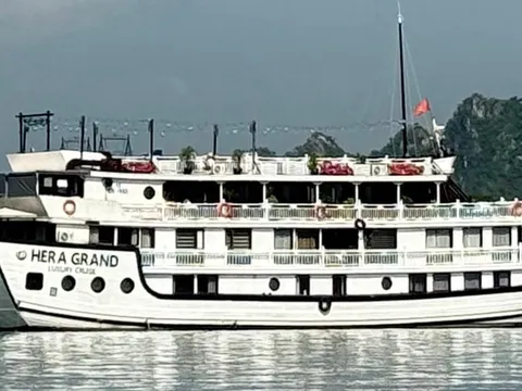 A white multi-deck wooden cruise boat with 'HERA GRAND Luxury Cruise' painted on its hull sits on calm water in front of green limestone karst hills, with other boats on either side.
