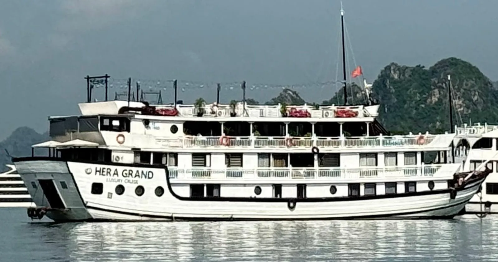 A white multi-deck wooden cruise boat with 'HERA GRAND Luxury Cruise' painted on its hull sits on calm water in front of green limestone karst hills, with other boats on either side.