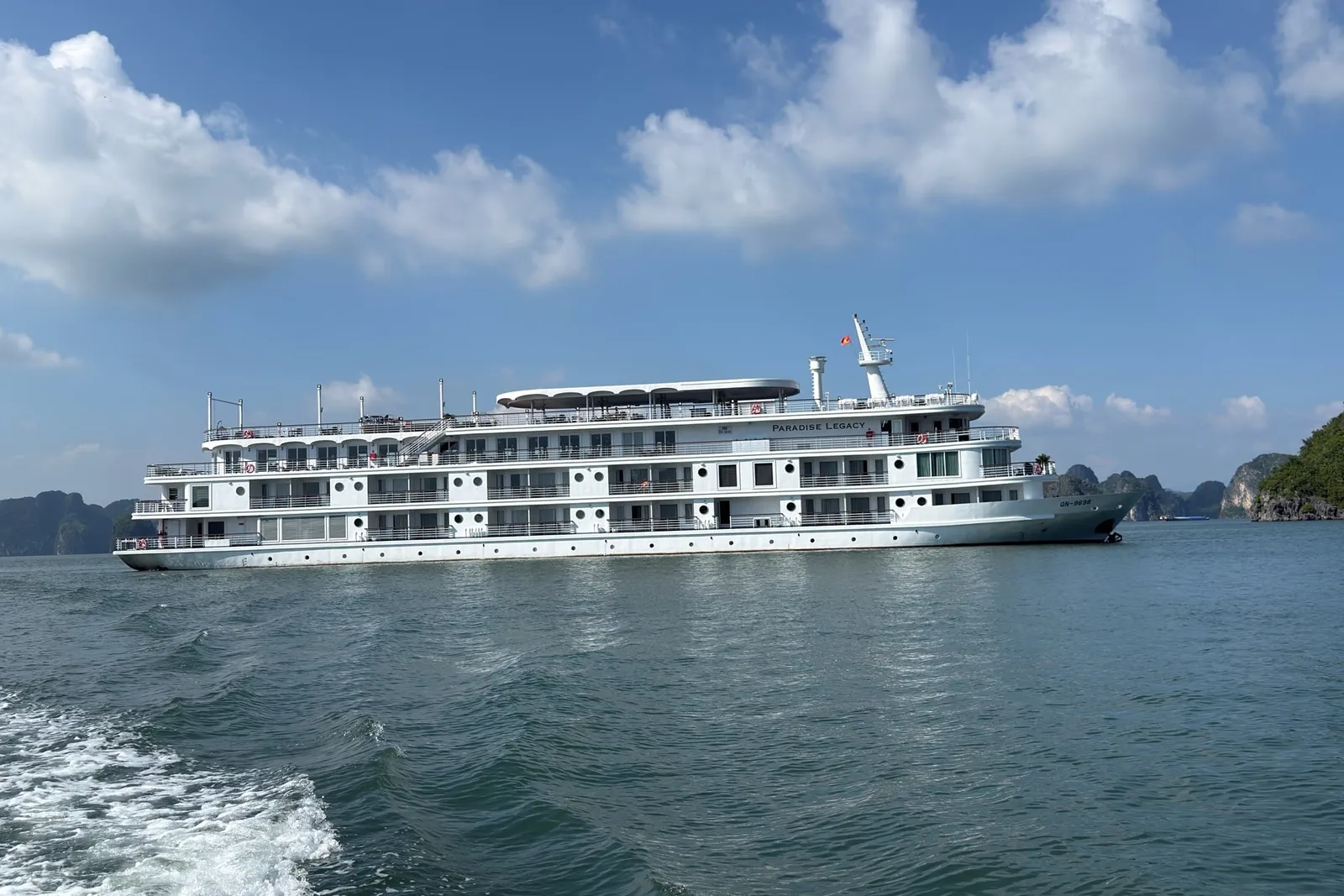 A large white multi-deck cruise ship with 'PARADISE LEGACY' lettered on its upper hull and QN-9698 on the bow, sailing on green water with limestone karst islands behind.