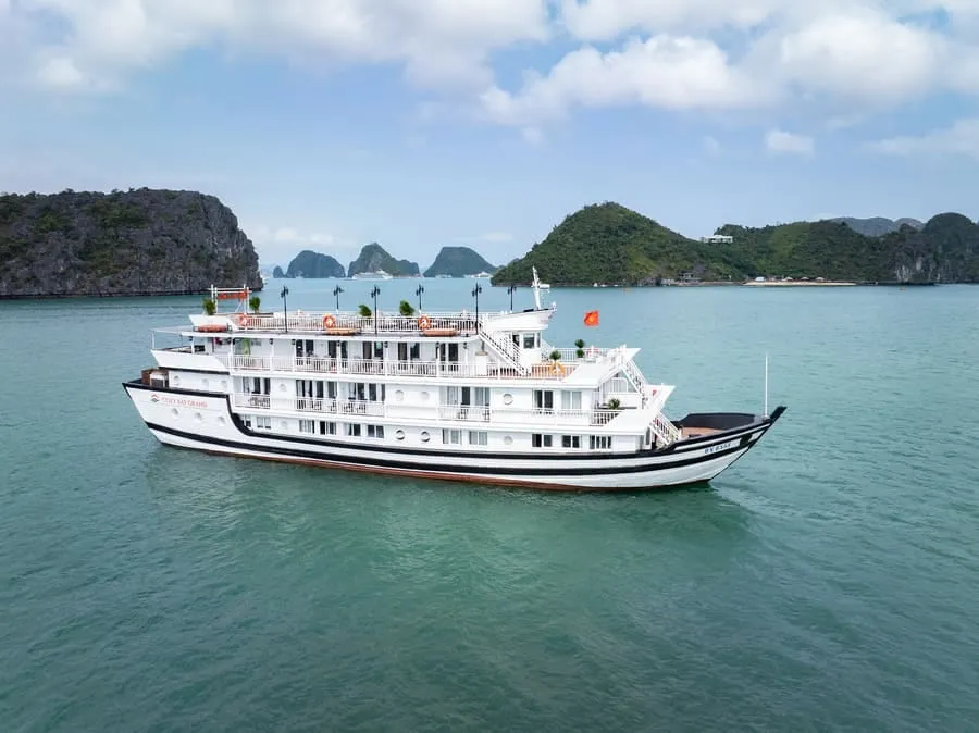 A white three-deck cruise boat with a black-and-white striped hull cruises on green water in front of karst islands under a blue sky.