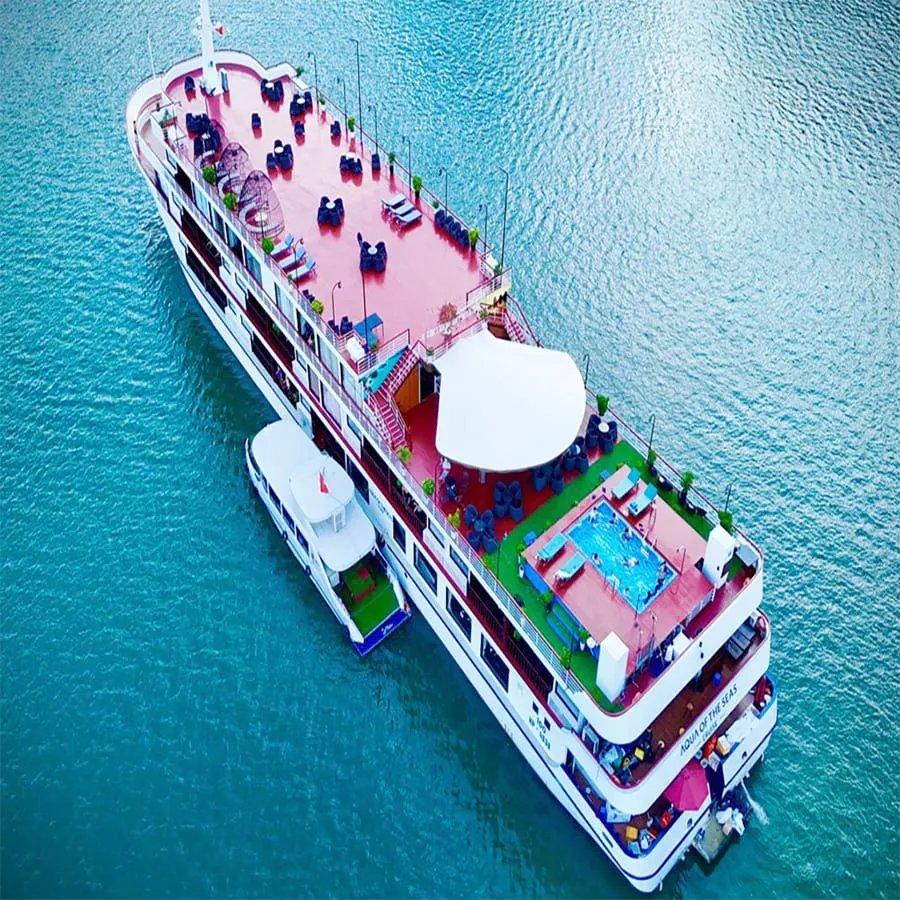 Aerial view of a white multi-deck cruise ship with pink deck areas, a swimming pool, and passengers seated at tables, floating in turquoise water.