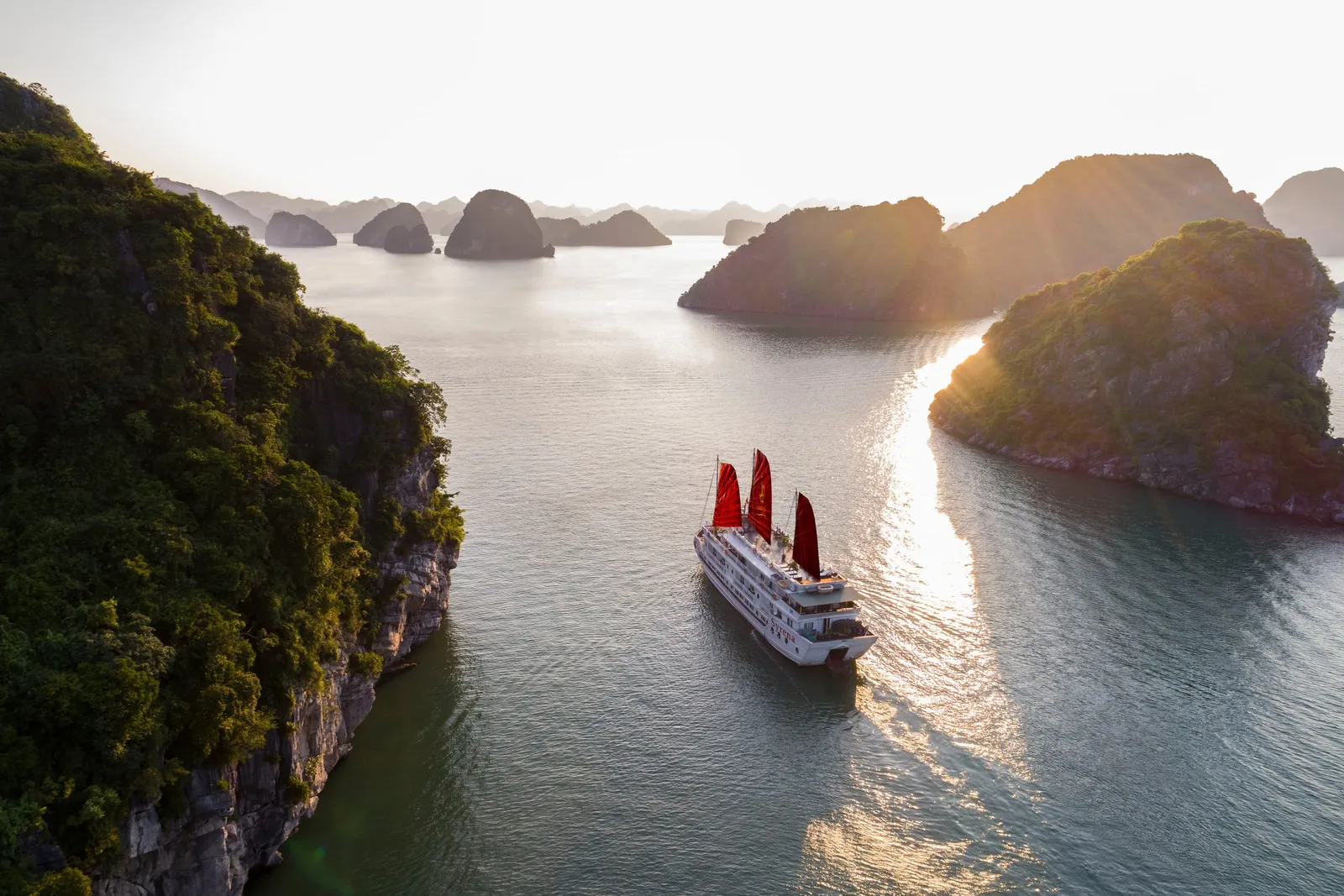 An aerial view of a white multi-deck cruise boat with three red sails moving through calm water between forested limestone karst islands in warm low sunlight.