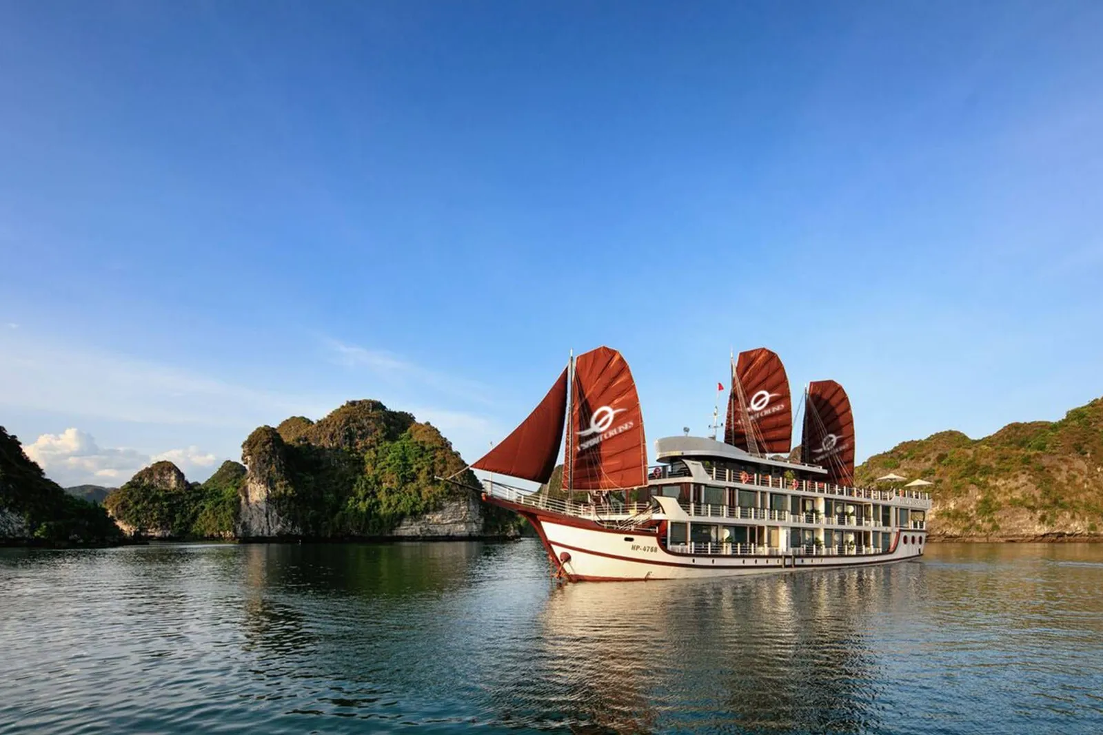 A multi-deck cruise boat with red-brown sails bearing a white logo and text sits on calm water beside green limestone karst islands under a blue sky.