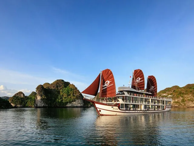 A multi-deck cruise boat with red-brown sails bearing a white logo and text sits on calm water beside green limestone karst islands under a blue sky.