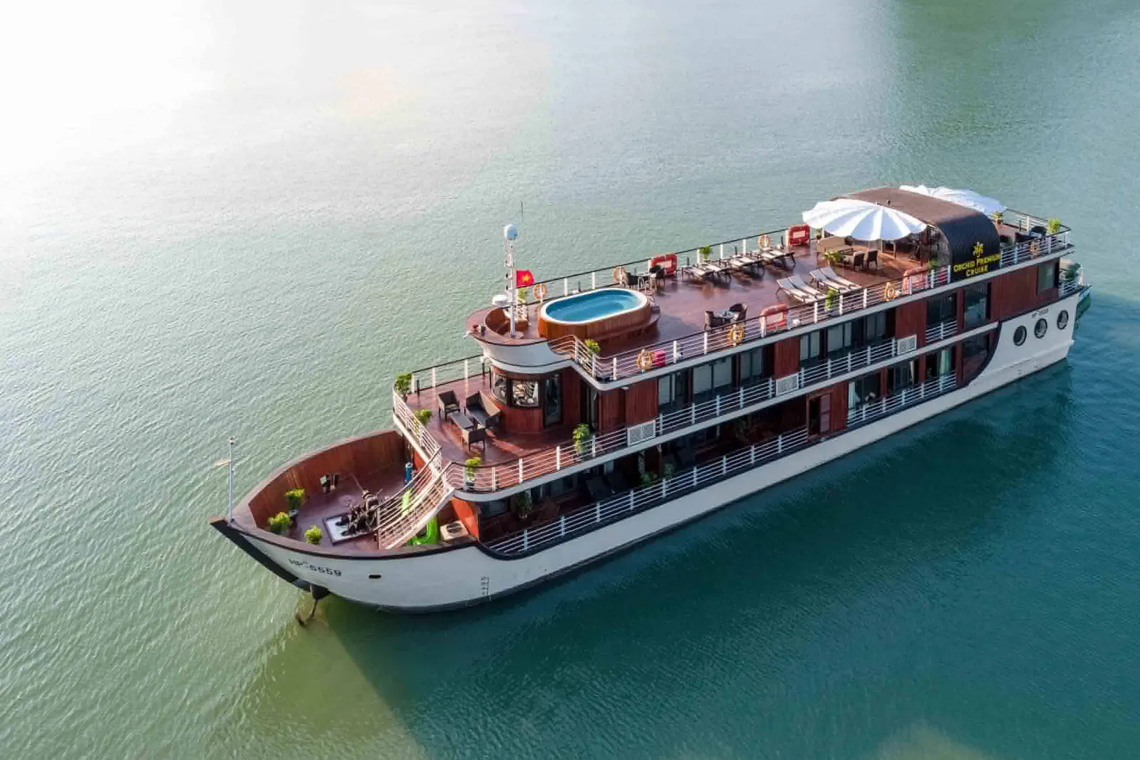 Aerial view of a multi-deck cruise ship with dark wood exterior, white hull, pool on top deck, and large umbrella, floating in calm turquoise water.