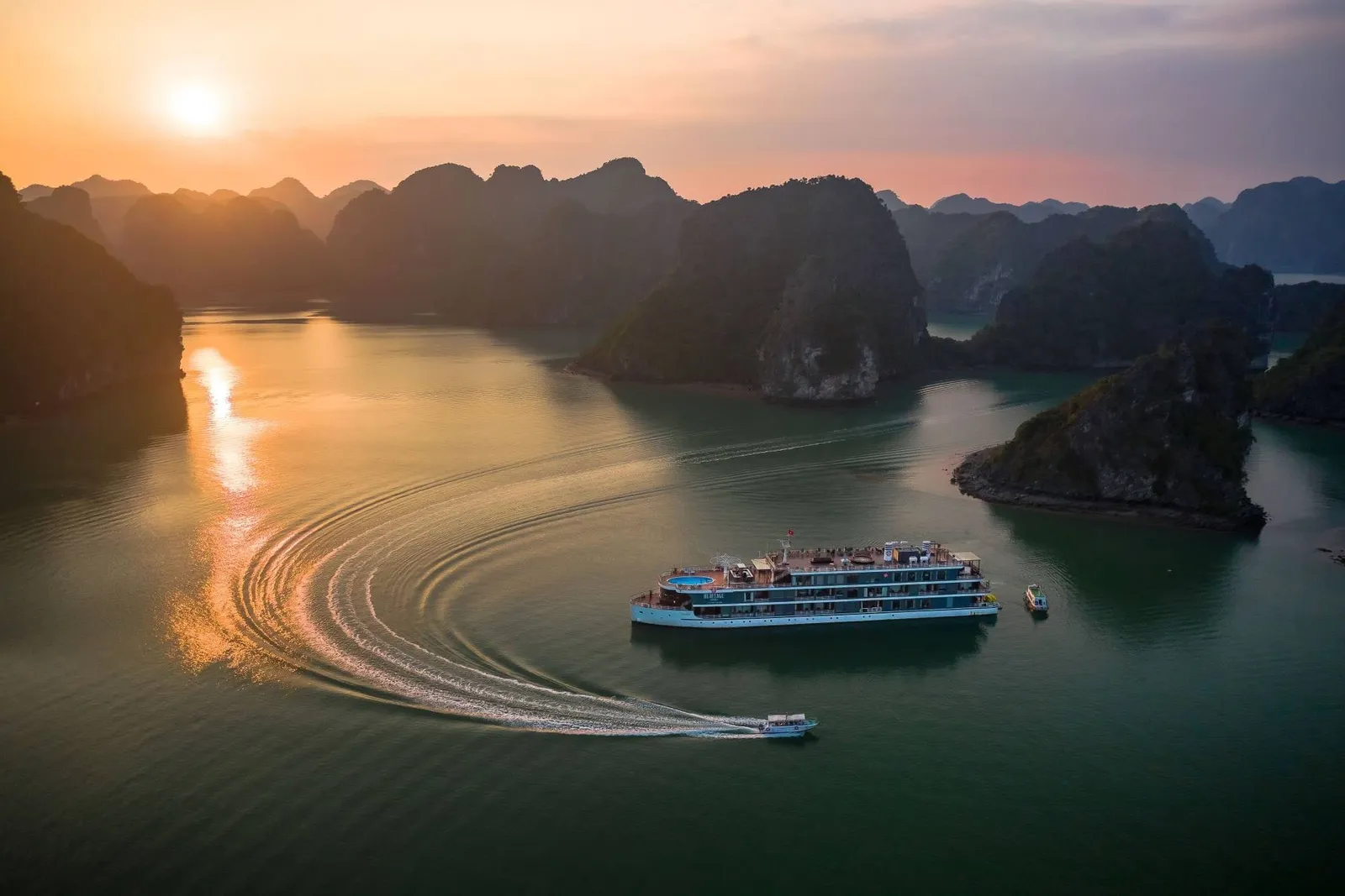 Aerial view at sunset of a multi-deck teal and white cruise ship with a rooftop pool among limestone karsts, with a small tender boat leaving a curved wake in the foreground.