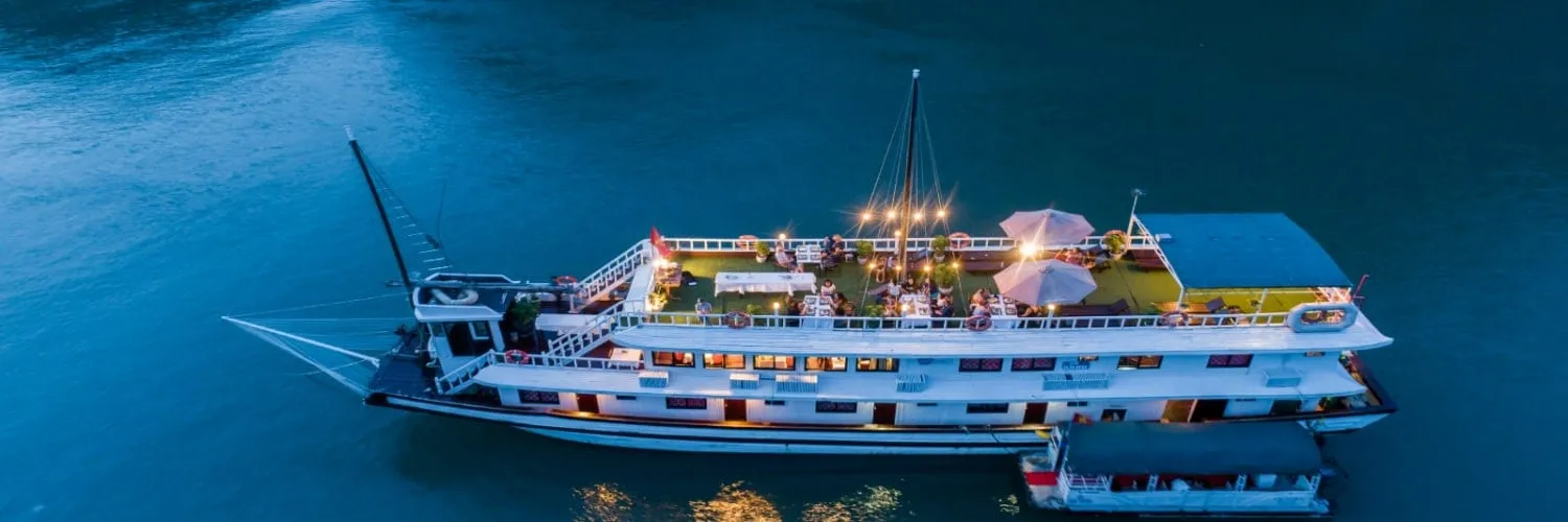 Aerial dusk view of a white multi-deck cruise boat with a lit sundeck, umbrellas, guests and a bare mast, with a smaller boat alongside on dark teal water.