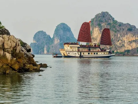 A cream junk with two red sails floating in front of tall limestone cliffs.