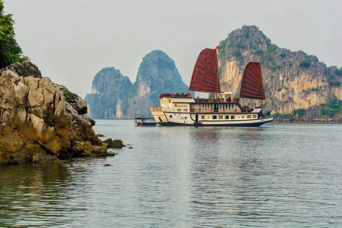 A cream junk with two red sails floating in front of tall limestone cliffs.