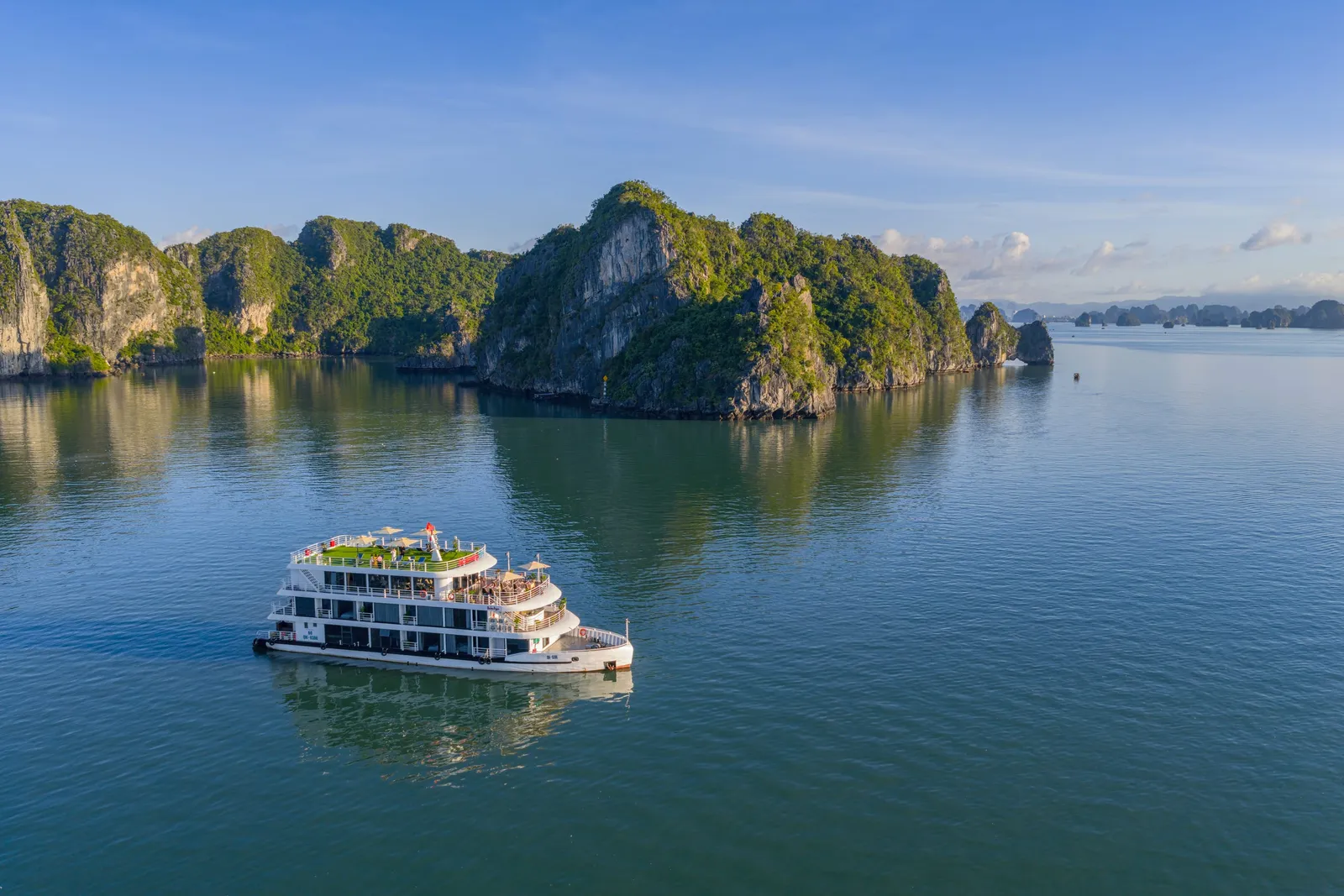 A white four-deck cruise boat sailing across calm water in front of forested limestone islands under a clear sky.