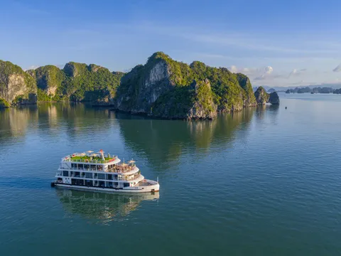 A white four-deck cruise boat sailing across calm water in front of forested limestone islands under a clear sky.