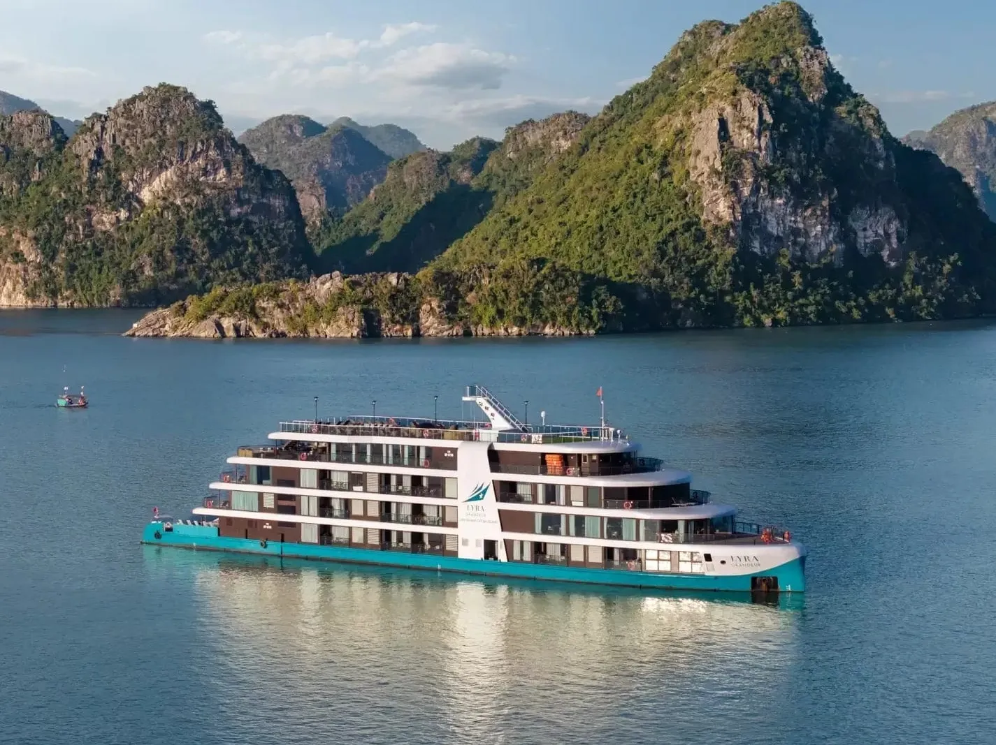 A modern white and teal cruise ship with multiple decks sails in calm water with dramatic limestone karst mountains in the background.