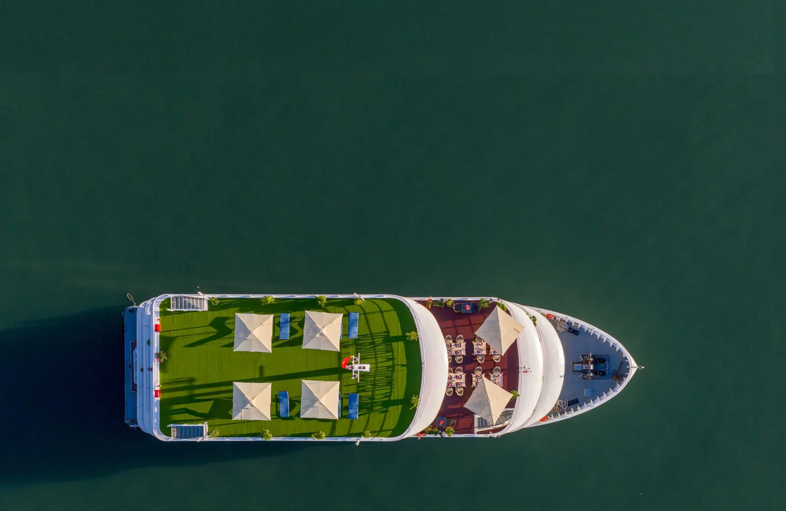A top-down aerial view of a white cruise boat with a green sundeck and white parasols on dark green water.
