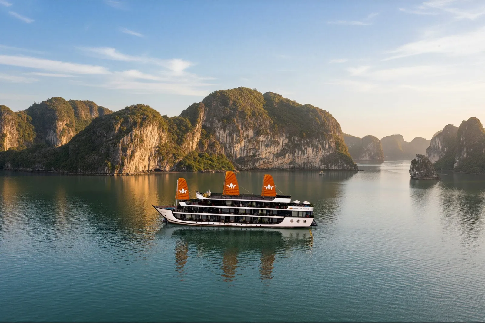 A white multi-deck cruise ship with three orange sails bearing logos floats in calm green water surrounded by limestone karst mountains.