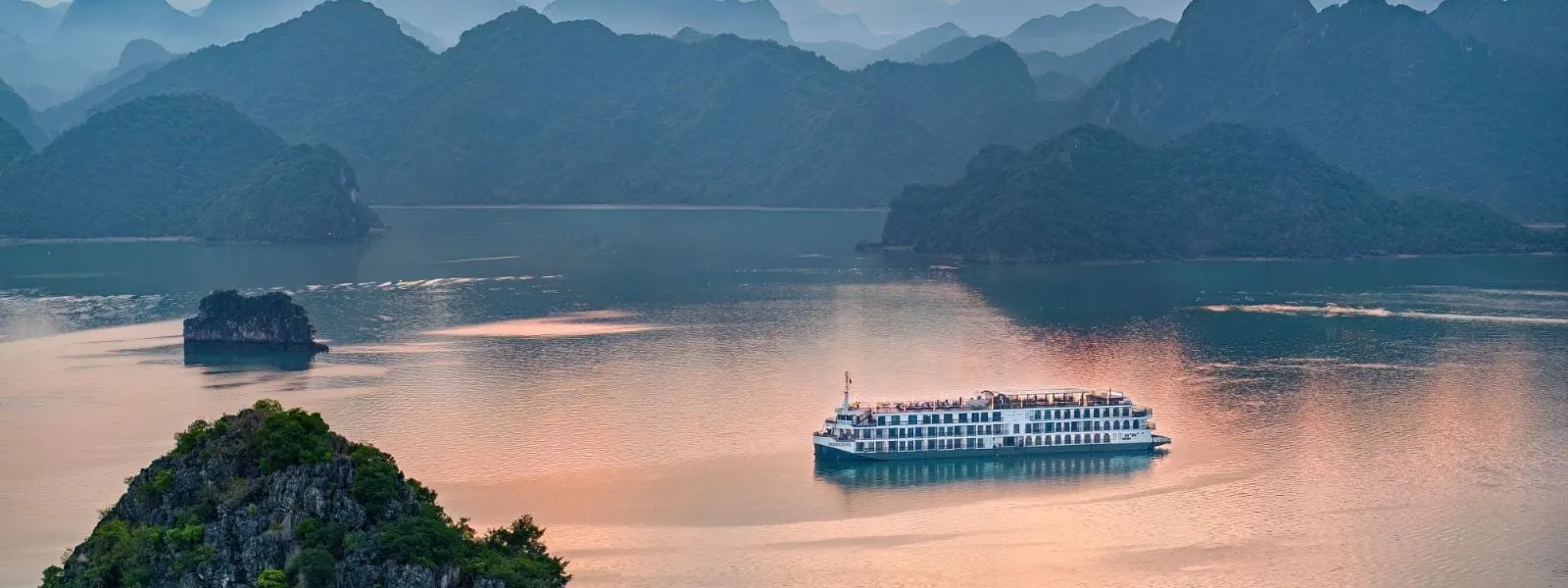 A white multi-deck steel cruise ship sits on calm pink-tinted water at dusk, seen from a high viewpoint, with forested limestone karsts around it.