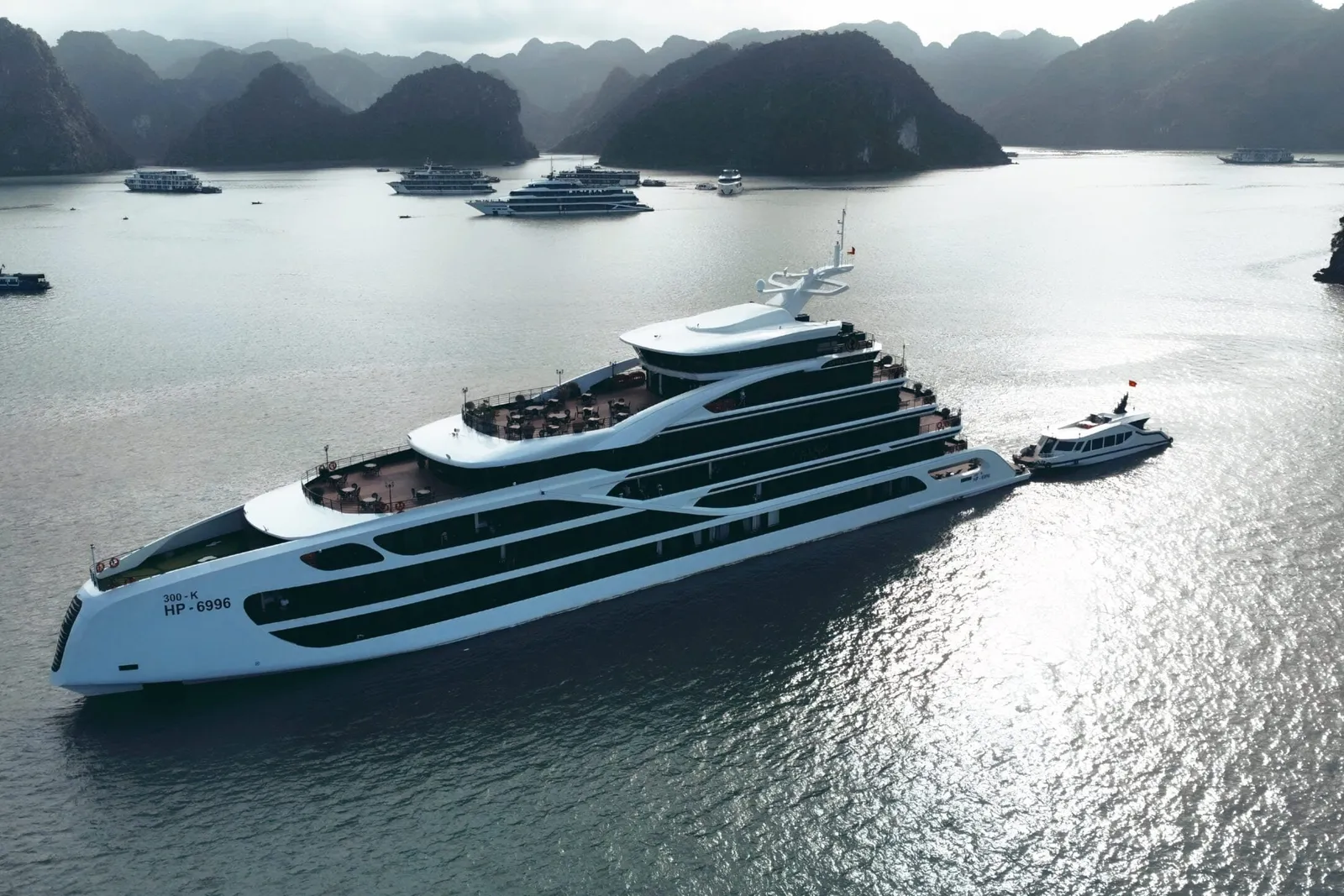 Aerial view of a large white multi-deck cruise ship with a small tender boat alongside, among limestone karst islands and other vessels on calm water.