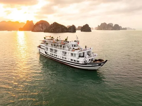 A white three-deck cruise boat at golden hour with the sun low over the water and limestone islands behind it.