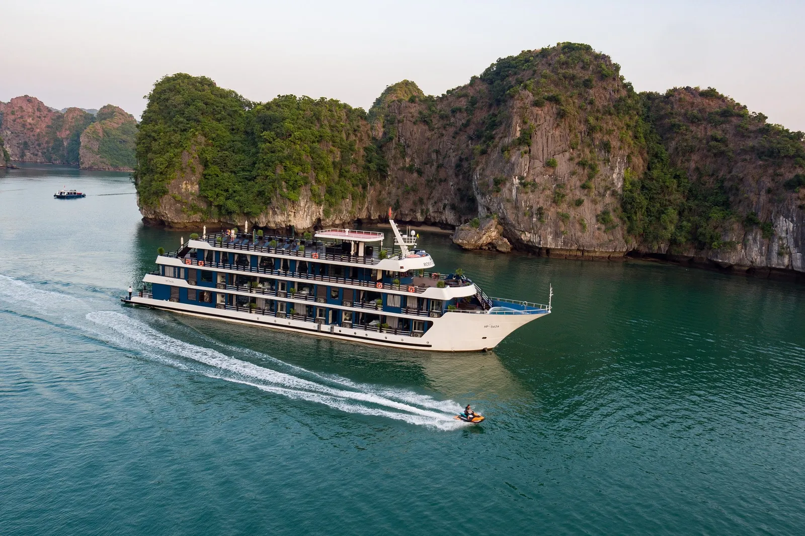 Aerial view of a white and blue multi-deck cruise ship sailing beside forested limestone karst cliffs, with a jet ski leaving a wake in the foreground.