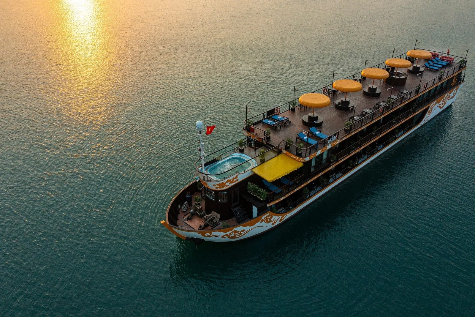 Aerial view of a multi-deck dark-hulled cruise ship with ornate gold decorative swirls, orange umbrellas and sun loungers on the top deck, a small plunge pool on the upper deck, and a Vietnamese flag, on calm water with a golden sunset reflection.