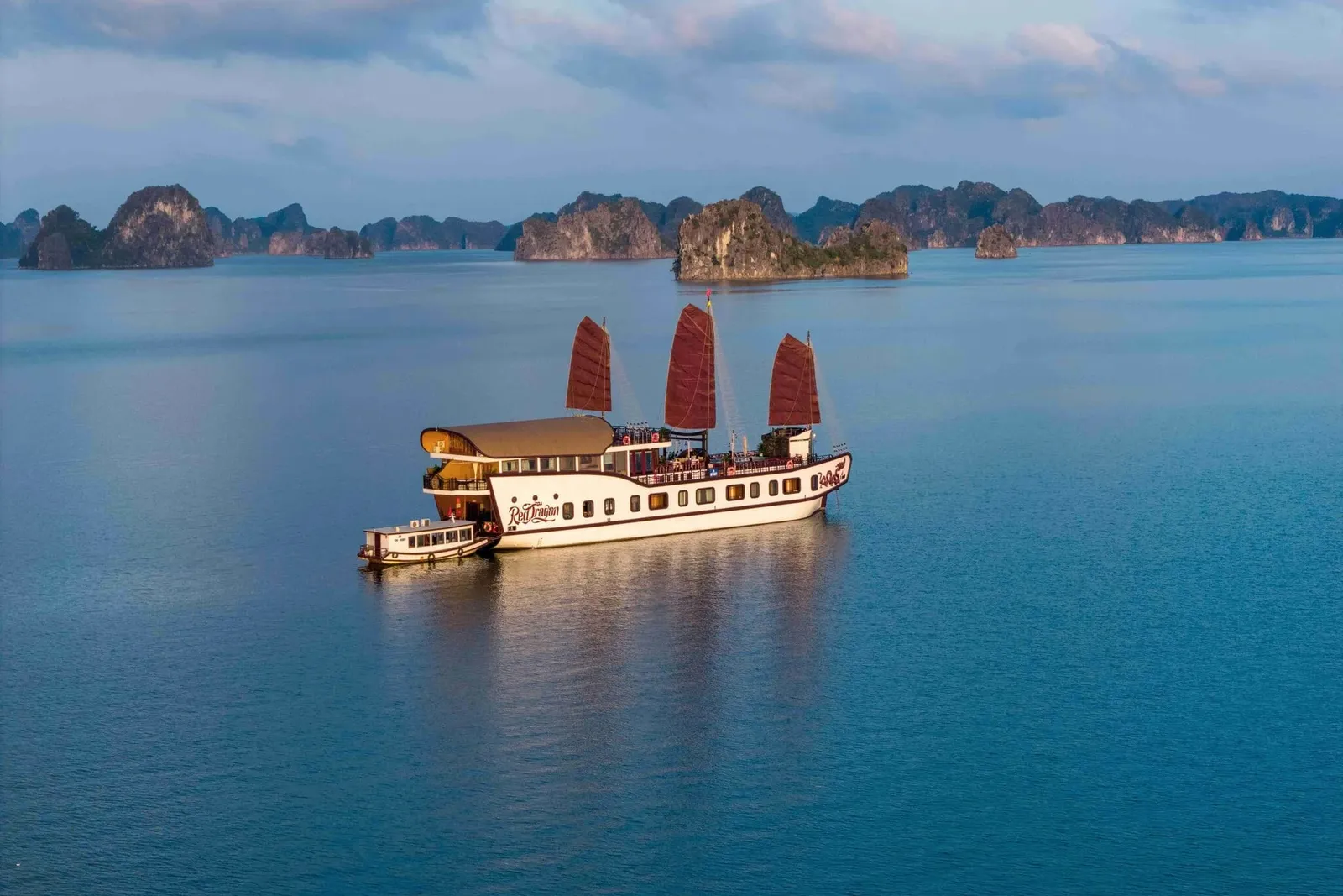 An aerial view of a white cruise junk with three red-brown sails and script lettering on its hull, anchored on calm blue water with a small tender alongside and limestone karst islands behind.