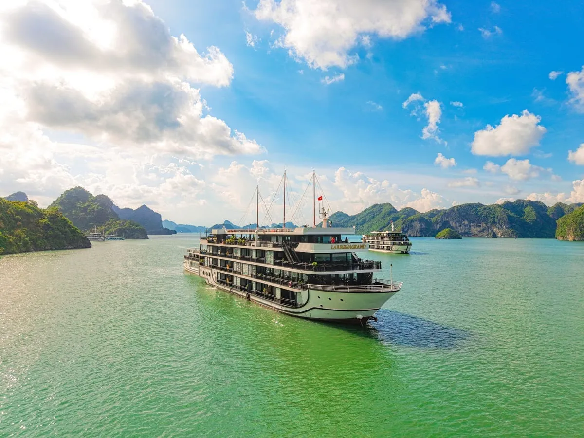 A multi-deck white cruise ship with a gold name sign on its upper deck sails on green water among forested limestone islands, with another vessel in the background and a blue sky with clouds.