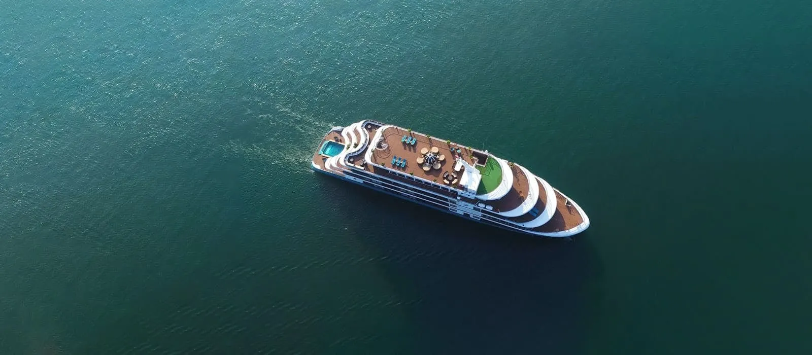 Aerial drone view of a white multi-deck cruise ship with a wooden top deck, small pool at the stern and a green patch at the bow, on calm dark teal water.