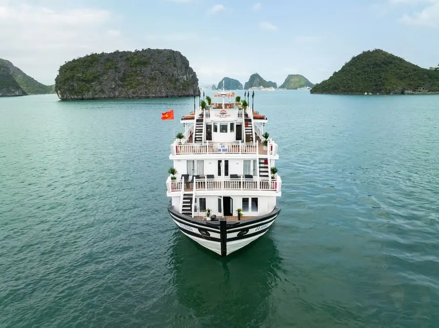 The bow of a white cruise boat with a striped hull, head-on, between two green karst islands.